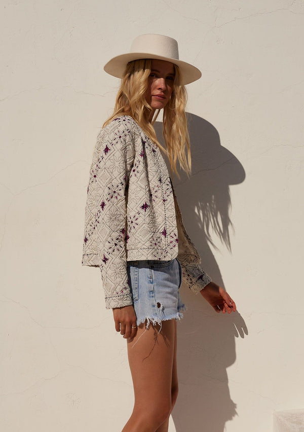 [Color: Natural/Fuchsia] A side facing image of a blonde model standing outside wearing a lightweight bohemian spring jacket in ivory with pink embroidered detail and contrast black thread detail. With long sleeves, an open front, and a cropped fit.
