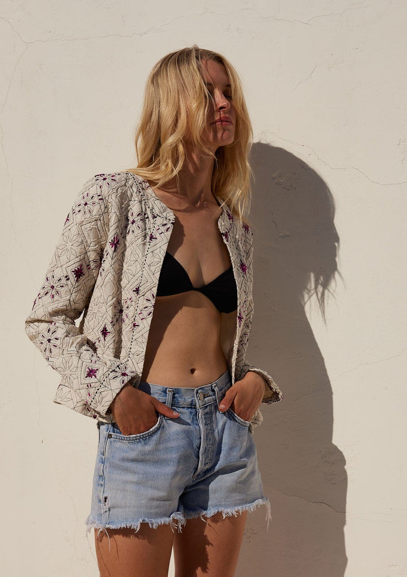 [Color: Natural/Fuchsia] A front facing image of a blonde model standing outside wearing a lightweight bohemian spring jacket in ivory with pink embroidered detail and contrast black thread detail. With long sleeves, an open front, and a cropped fit.