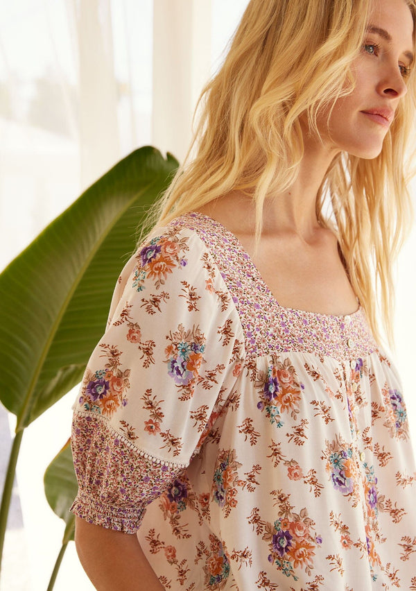 [Color: Natural/Purple] A close up side facing image of a blonde model wearing a bohemian resort blouse in an off white and purple floral border print. With short puff sleeves, a square neckline, a button front, an adjustable drawstring waist, and a mini pom trim.