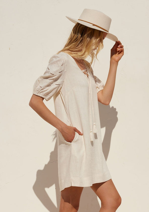 [Color: Vanilla] A half body side facing image of a blonde model wearing an off white bohemian mini dress with short puff sleeves, embroidered detail, side pockets, a v neckline with lace up detail and tassel ties, and a relaxed, loose fit.
