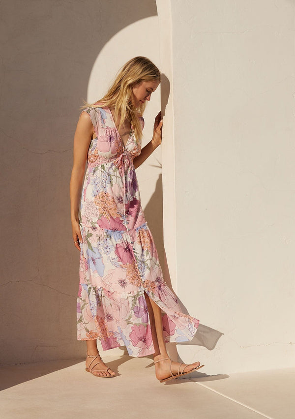 [Color: Ivory/Natural] A side facing image of a blonde model standing outside wearing a pink floral maxi dress. With short cap sleeves, a v neckline, a button front, an empire waist, a ruffle trimmed tiered skirt, and a drawstring waist with tassel ties.