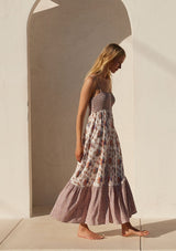 [Color: Natural/Purple] A side facing image of a blonde model wearing a best selling bohemian maxi dress in an off white and purple floral border print. With adjustable spaghetti straps, a square neckline, a smocked fitted bodice, a flowy tiered skirt, side pockets, and a button front.