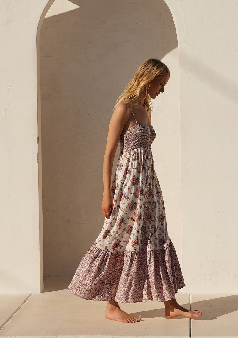 [Color: Natural/Purple] A side facing image of a blonde model wearing a best selling bohemian maxi dress in an off white and purple floral border print. With adjustable spaghetti straps, a square neckline, a smocked fitted bodice, a flowy tiered skirt, side pockets, and a button front.