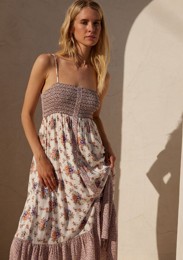 [Color: Natural/Purple] A half body front facing image of a blonde model standing outside wearing a best selling bohemian maxi dress in an off white and purple floral border print. With adjustable spaghetti straps, a square neckline, a smocked fitted bodice, a flowy tiered skirt, side pockets, and a button front.