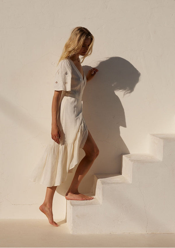 [Color: Vanilla] A side facing image of a blonde model standing outside wearing an off white bohemian mid length wrap dress. With short flutter sleeves, embroidered detail, a deep v neckline, a side tie waist closure, and a flowy tiered skirt.