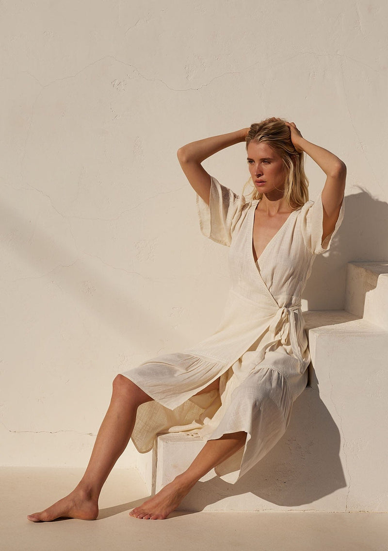 [Color: Vanilla] A front facing image of a blonde model sitting outside wearing an off white bohemian mid length wrap dress. With short flutter sleeves, embroidered detail, a deep v neckline, a side tie waist closure, and a flowy tiered skirt.