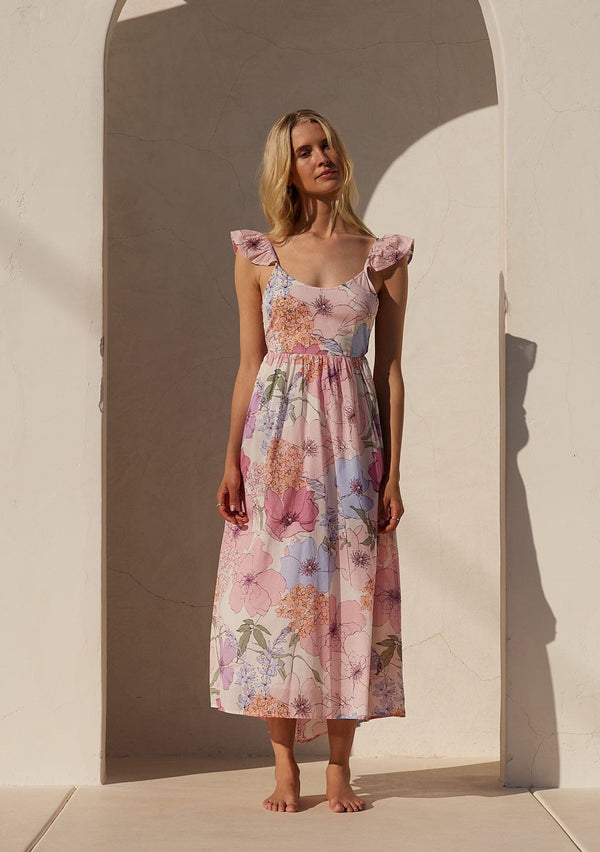 [Color: Ivory/Natural] A front facing image of a blonde model standing outside wearing a pink floral maxi dress. With short flutter cap sleeves, a scoop neckline, a cutout back detail with adjustable tie, a slim fit top, and a long flowy skirt.