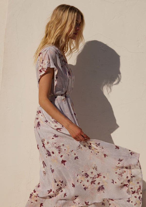 [Color: Dusty Blush/Wine] A close up side facing image of a blonde model standing outside wearing a bohemian maxi dress designed in a pink floral print and crafted from textured chiffon. With short dolman sleeves, a surplice v neckline, an open back with tie closure, and a flowy asymmetric hemline skirt.