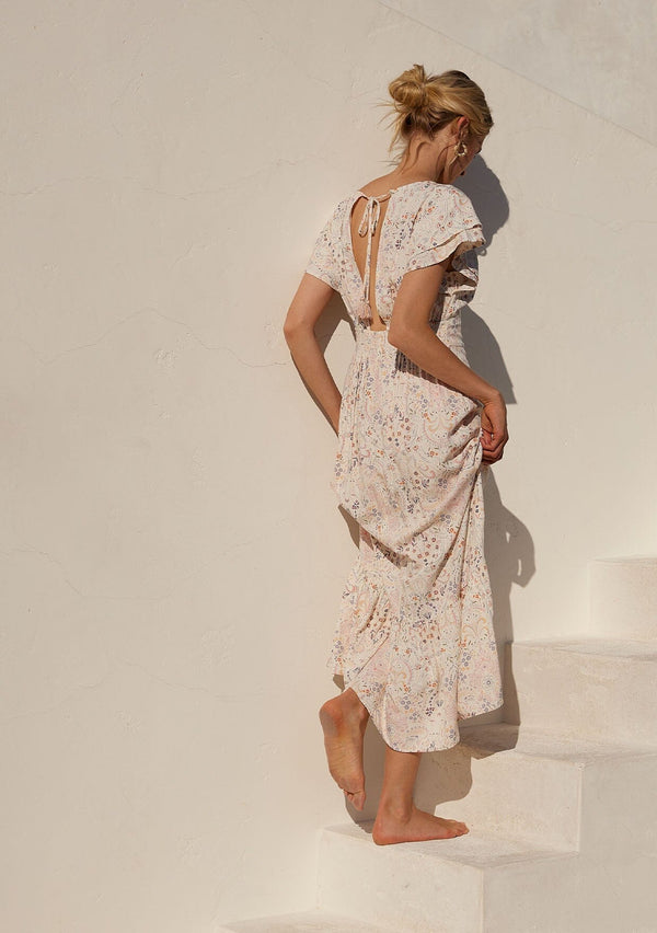 [Color: Natural/Peach] A back facing image of a blonde model standing outside wearing a bohemian special occasion maxi dress in a natural and pink floral print. With short flutter sleeves, a v neckline, a tiered skirt, an open back with tassel tie detail, and a half smocked elastic waist at the back. 