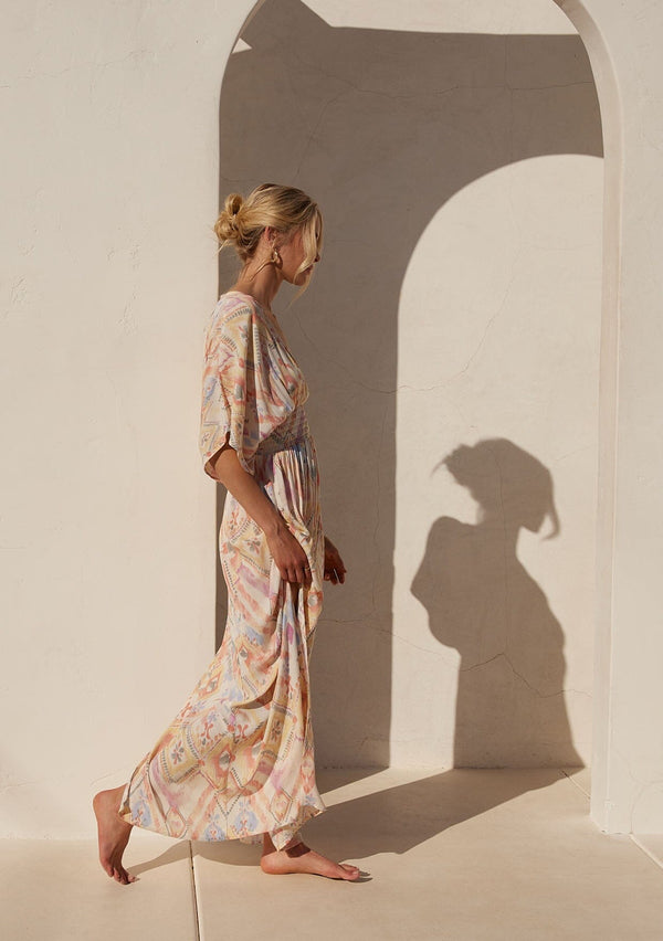 [Color: Butter/Coral] A side facing image of a blonde model standing outside wearing a best selling bohemian resort maxi dress in a yellow bohemian diamond print. With half length kimono sleeves, a deep v neckline in the front and back, a back tie closure, a smocked elastic waist, side slits, and a long flowy skirt.