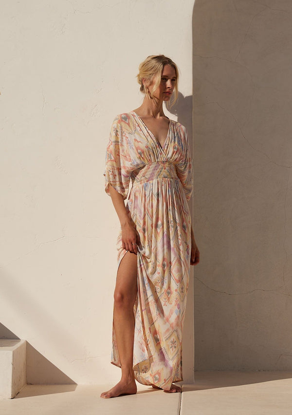 [Color: Butter/Coral] A front facing image of a blonde model standing outside wearing a best selling bohemian resort maxi dress in a yellow bohemian diamond print. With half length kimono sleeves, a deep v neckline in the front and back, a back tie closure, a smocked elastic waist, side slits, and a long flowy skirt.