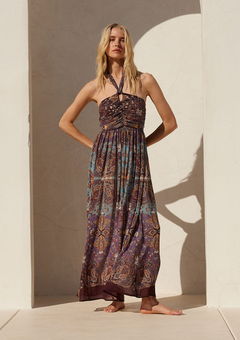[Color: Raisin/Teal] A full body front facing image of a blonde model standing outside wearing a bohemian halter maxi dress in a multi colored purple and teal floral print. With a tie neckline, a slim fit bodice with a smocked elastic back, and a long flowy skirt.