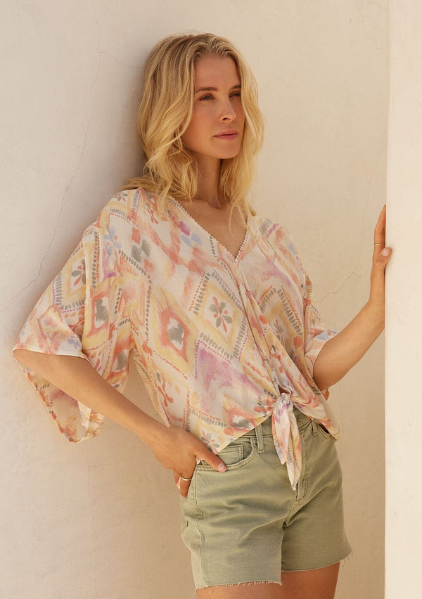[Color: Butter/Coral] A front facing image of a blonde model standing outside wearing a lightweight resort top designed in a yellow and coral bohemian diamond print. With half length wide sleeves, a v neckline, a tie front waist, and a mini pom trim.