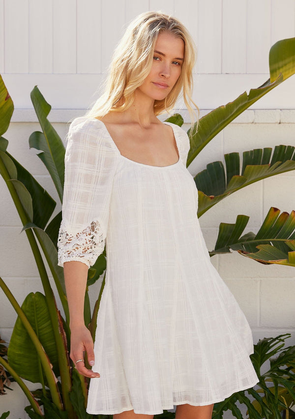 [Color: Off White] A front facing image of a blonde model standing outside wearing a white cotton bohemian mini dress in a textured dobby plaid fabric. With a flared paneled mini skirt, a scooped neckline, three quarter length sleeves with a crochet detail, and a relaxed fit. 