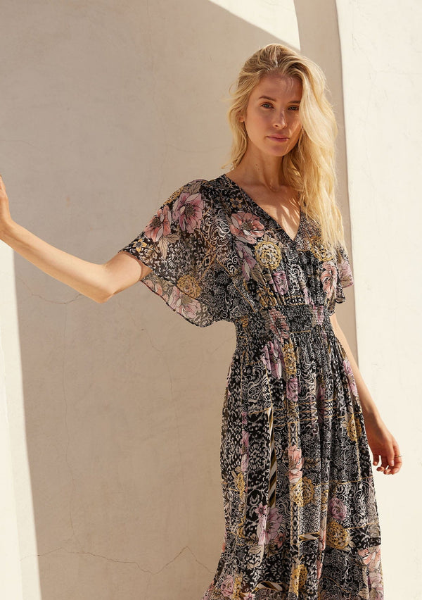 [Color: Black/Dusty Peach] A half body front facing image of a blonde model standing outside wearing a bohemian chiffon maxi dress designed in a black and pink mixed floral print. With short flutter sleeves, a v neckline, a smocked elastic waist, and a long flowy skirt with a tiered asymmetric hemline.