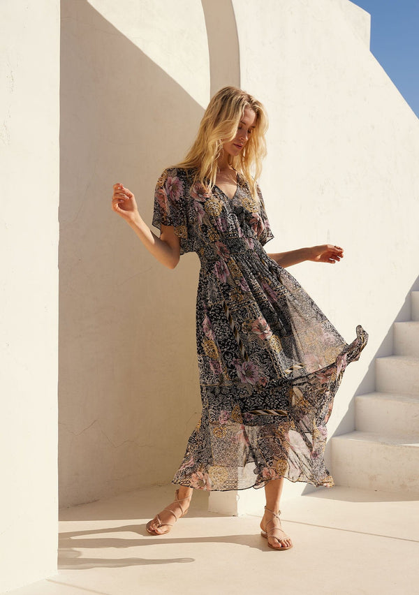[Color: Black/Dusty Peach] A front facing image of a blonde model standing outside wearing a bohemian chiffon maxi dress designed in a black and pink mixed floral print. With short flutter sleeves, a v neckline, a smocked elastic waist, and a long flowy skirt with a tiered asymmetric hemline.