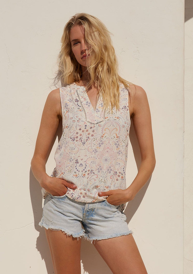 [Color: Natural/Pink] A front facing image of a blonde model standing in the sun wearing a relaxed bohemian resort tank top in a natural and pink floral print. With a v neckline, sheer lattice trim, and a soft hand feel.