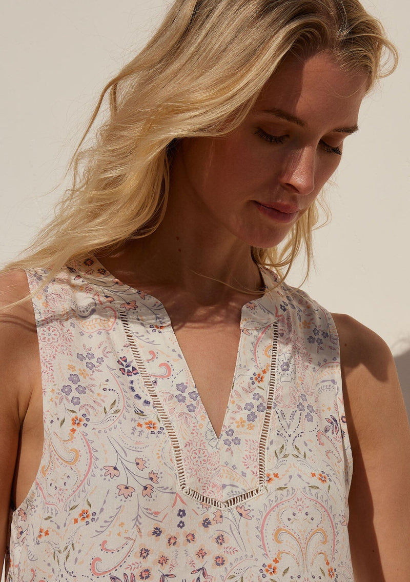 [Color: Natural/Pink] A close up front facing image of a blonde model standing in the sun wearing a relaxed bohemian resort tank top in a natural and pink floral print. With a v neckline, sheer lattice trim, and a soft hand feel.