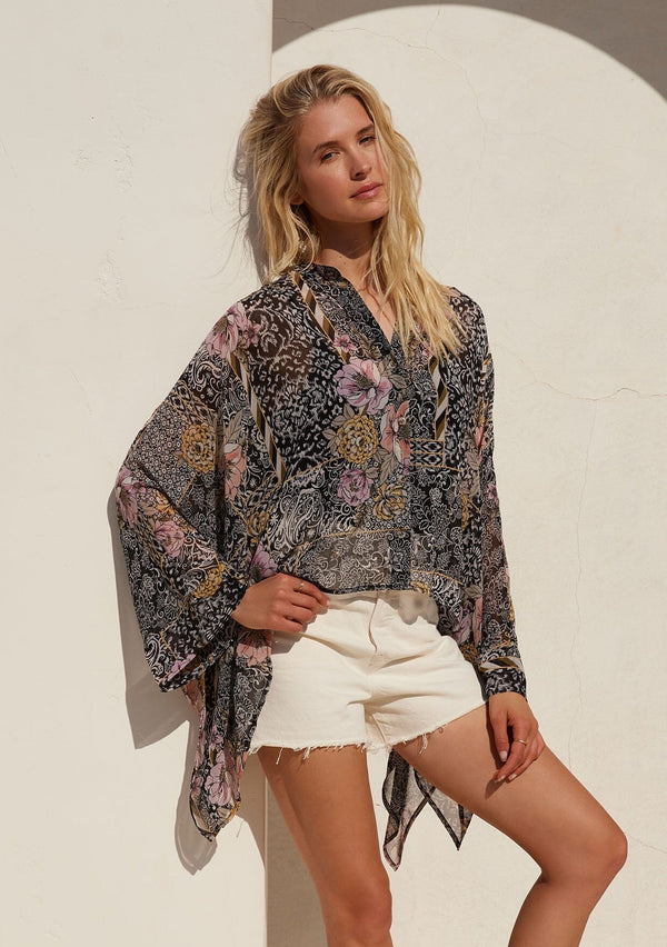 [Color: Black/Dusty Peach] A front facing image of a blonde model standing outside wearing an ultra flowy bohemian resort blouse in a black and pink mixed floral print. Crafted from chiffon and featuring long dolman sleeves, a high round neckline, side slits, and a button front.