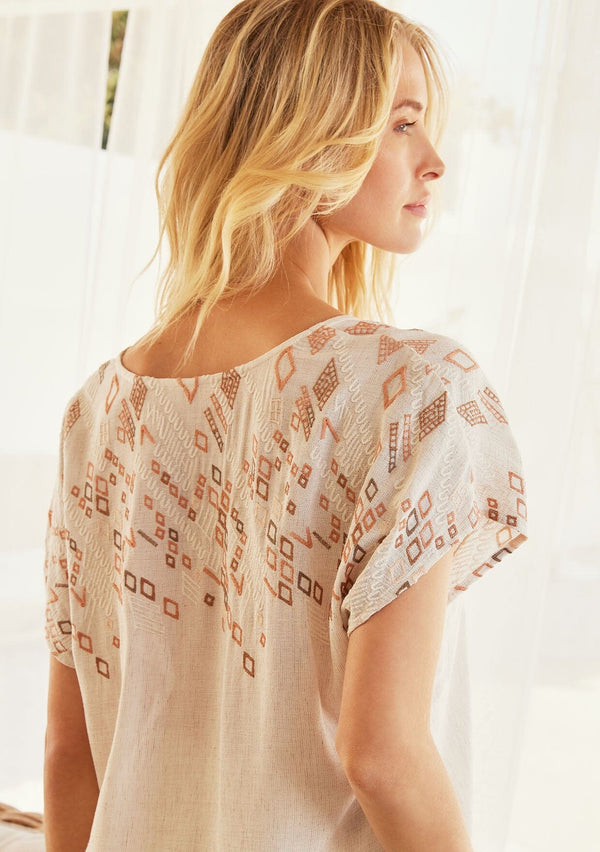 [Color: Natural/Hazel] A close up back facing image pf a blonde model standing outside wearing an ivory boxy fit bohemian resort top with brown embroidered detail. With short dolman sleeves, a split v neckline, and tassel ties.
