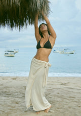 [Color: Vanilla] A front facing image of a brunette model wearing an off-white linen blend wrap skirt with a side tie and smocked back, styled for warm weather, vacation, or beach days.