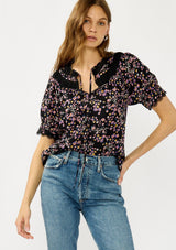 [Color: Black/Pink] A front facing image of a black and pink floral print top with puff sleeves, a split v-neckline with ties, shirred details, and lace trims. A day to night versatile top for the fall season. 