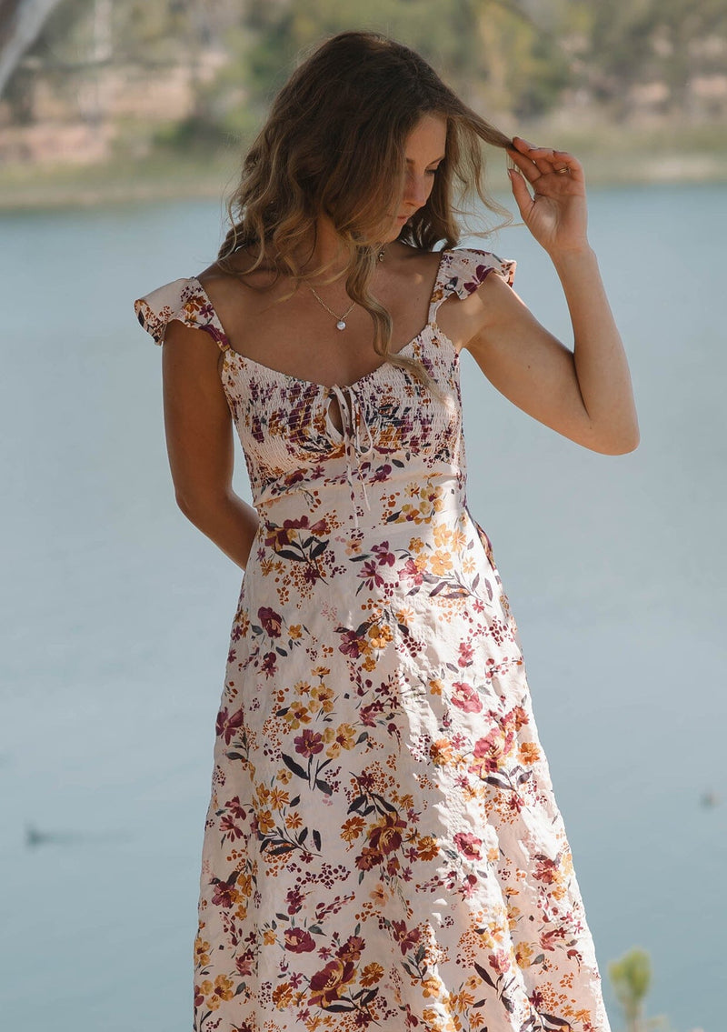 [Color: Almond/Wine] A front facing image of a blonde model wearing a pink floral print midi dress with a scooped neckline, smocked bust and back, elastic shoulder straps with ruffle trim, and sexy cutout back detail. 