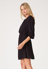 [Color: Black] A side facing image of a blonde model wearing a classic bohemian black mini dress with three quarter length sleeves, a self covered button front, an elastic waist, and a pleated round neckline.