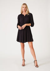 [Color: Black] A full body front facing image of a blonde model wearing a classic bohemian black mini dress with three quarter length sleeves, a self covered button front, an elastic waist, and a pleated round neckline.
