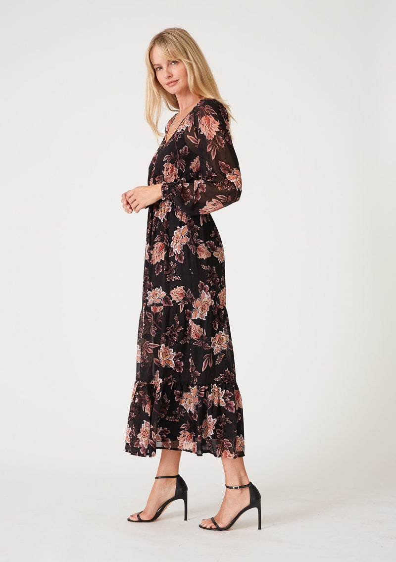 [Color: Black/Dusty Coral] A side facing image of a blonde model wearing a bohemian black and coral floral print chiffon maxi dress. With voluminous long sleeves, a v neckline, a flowy tiered skirt, and an elastic waist.