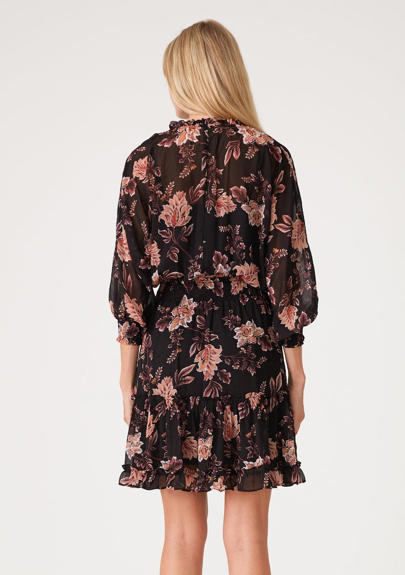 [Color: Black/Dusty Coral] A back facing image of a blonde model wearing a black chiffon bohemian mini dress with a coral pink floral print throughout. With voluminous long sleeves, a split v neckline with ties, ruffled trim throughout, and a smocked elastic waist for added definition.