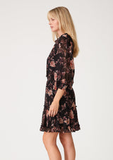 [Color: Black/Dusty Coral] A side facing image of a blonde model wearing a black chiffon bohemian mini dress with a coral pink floral print throughout. With voluminous long sleeves, a split v neckline with ties, ruffled trim throughout, and a smocked elastic waist for added definition.
