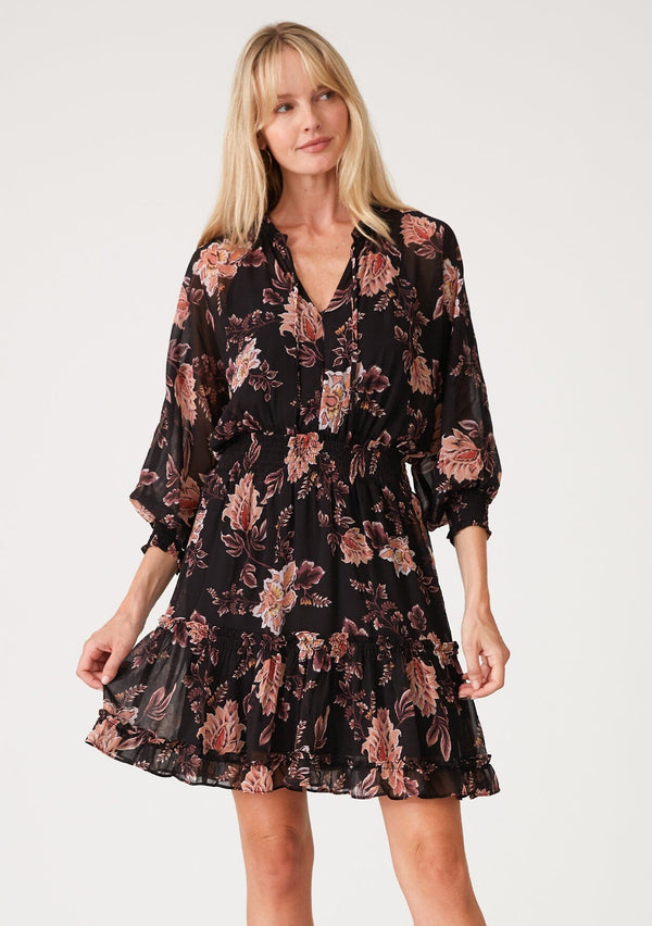 [Color: Black/Dusty Coral] A front facing image of a blonde model wearing a black chiffon bohemian mini dress with a coral pink floral print throughout. With voluminous long sleeves, a split v neckline with ties, ruffled trim throughout, and a smocked elastic waist for added definition.