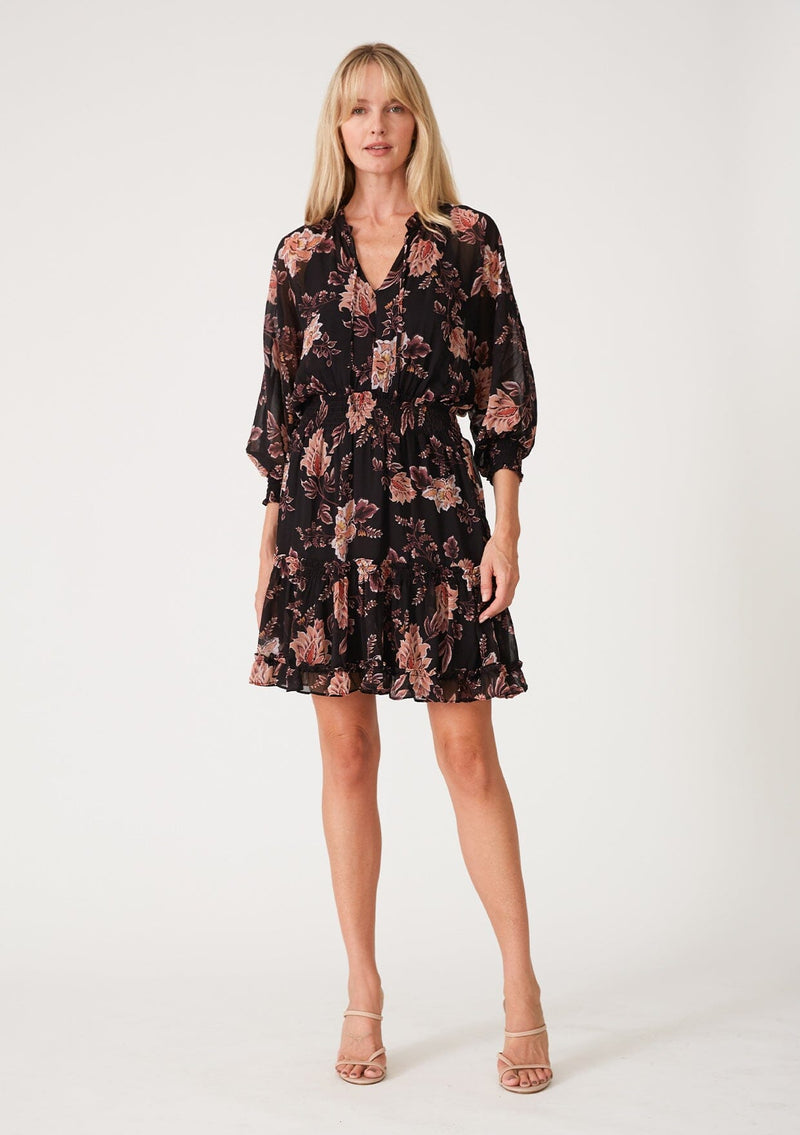 [Color: Black/Dusty Coral] A full body front facing image of a blonde model wearing a black chiffon bohemian mini dress with a coral pink floral print throughout. With voluminous long sleeves, a split v neckline with ties, ruffled trim throughout, and a smocked elastic waist for added definition.