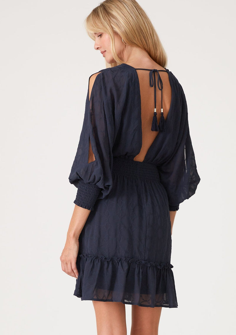 [Color: Ink Blue] A back facing image of a blonde model wearing a navy blue bohemian mini dress in embroidered chiffon. With long split sleeves, a ruffle trimmed tiered skirt, a smocked elastic waist, a v neckline, and an open back with tassel tie closure. Perfect for weddings or date nights.