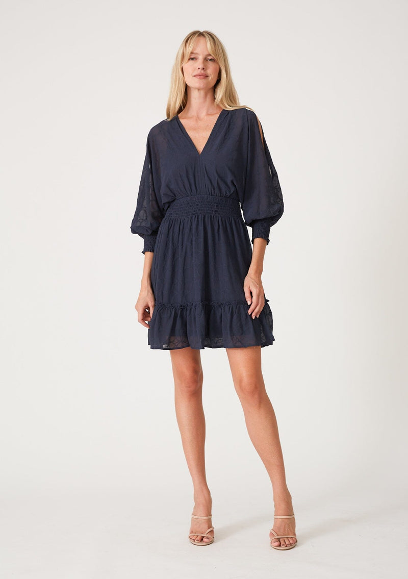 [Color: Ink Blue] A full body front facing image of a blonde model wearing a navy blue bohemian mini dress in embroidered chiffon. With long split sleeves, a ruffle trimmed tiered skirt, a smocked elastic waist, a v neckline, and an open back with tassel tie closure. Perfect for weddings or date nights.