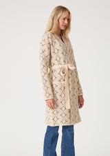 [Color: Natural/Taupe] A side facing image of a blonde model wearing a soft mid length cardigan sweater coat in an ivory diamond jacquard. With long sleeves, side pockets, and a belted tie waist.