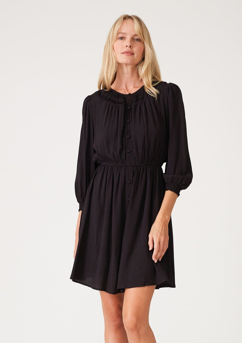 [Color: Black] A front facing image of a blonde model wearing a classic bohemian black mini dress with three quarter length sleeves, a self covered button front, an elastic waist, and a pleated round neckline.