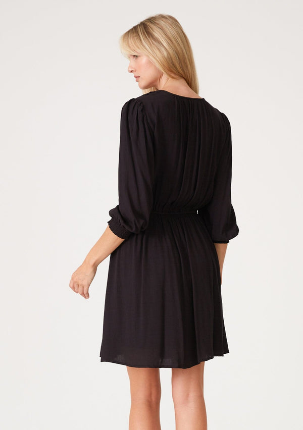 [Color: Black] A back facing image of a blonde model wearing a classic bohemian black mini dress with three quarter length sleeves, a self covered button front, an elastic waist, and a pleated round neckline.