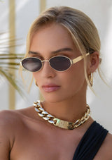 [Color: Brown] A woman wearing thin gold metal sunglasses with an oval shape and dark tan lenses. Made by FREYRS