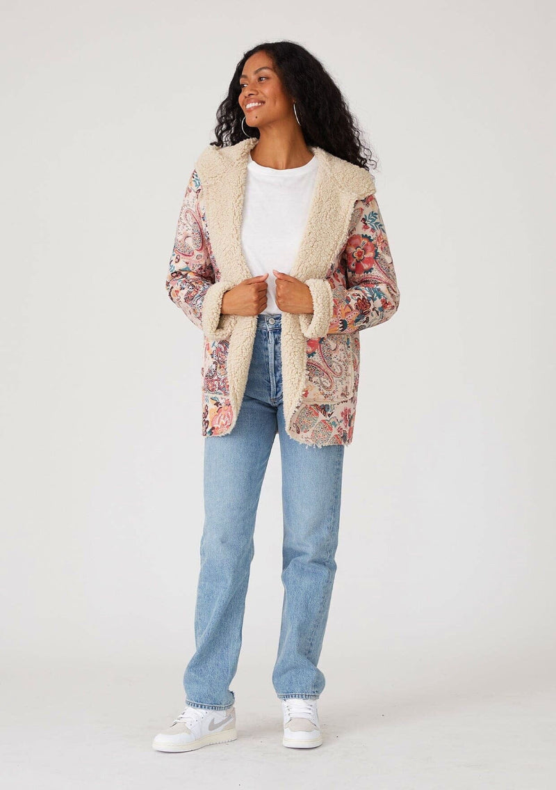 [Color: Natural/Red] A front facing image of a brunette model wearing a bohemian jacket with a pink and natural floral print and a faux shearling lining. With long sleeves, side pockets, a hoodie, and an open front.
