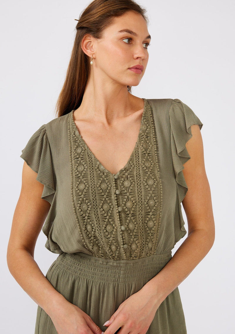 [Color: Olive] A close up front facing image of a brunette model wearing an olive green bohemian short romper. With short flutter sleeves, a v neckline, a crochet top, a self covered button front top, and a smocked elastic waist. 