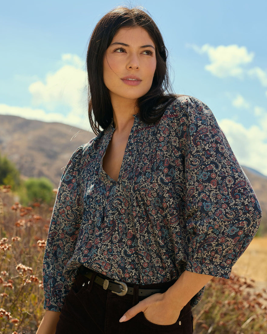 Woman in a patterned blouse standing outdoors with mountains in the background