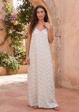 [Color: Aqua/Peach] A woman modeling a sleeveless, floor-length maxi dress with an abstract print and a zigzag pattern standing in a sunlit courtyard with terracotta tiles and a warm stucco wall.