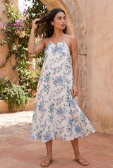 [Color: Ocean/Blue] A brunette model standing near greenery and a stone wall wearing a blue and white halter maxi dress with a relaxed, flowy silhouette. 