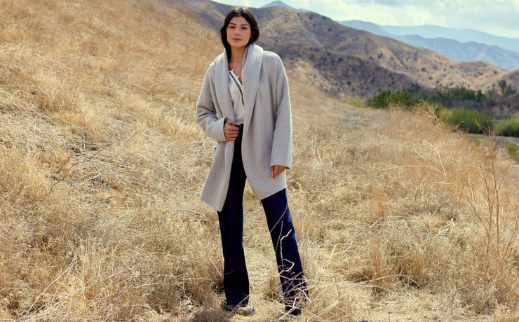 Woman in a long coat standing in a dry, open landscape with mountains in the background
