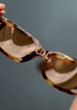 [Color: Tortie] A classic rounded rectangular frame in a brown tortoiseshell frame, made with biodegradable acetate. With polarized lenses and the word fearless on the inner frame. 