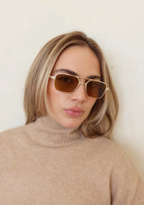 [Color: Brown] A woman wearing a  matte gold metal straight frame sunglasses with 100% UVA/UVB protected brown lenses. 