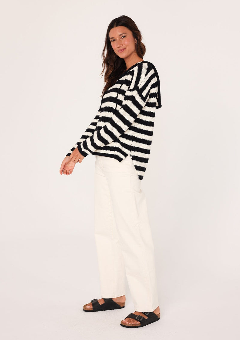 [Color: Black/White] A side facing image of a brunette model wearing a black and white striped knit hoodie. With long sleeves, a dropped shoulder, a v neckline, and a front kangaroo pocket. 