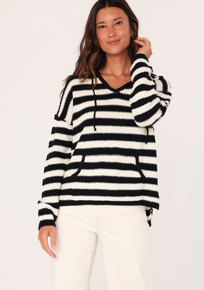 [Color: Black/White] A front facing image of a brunette model wearing a black and white striped knit hoodie. With long sleeves, a dropped shoulder, a v neckline, and a front kangaroo pocket. 